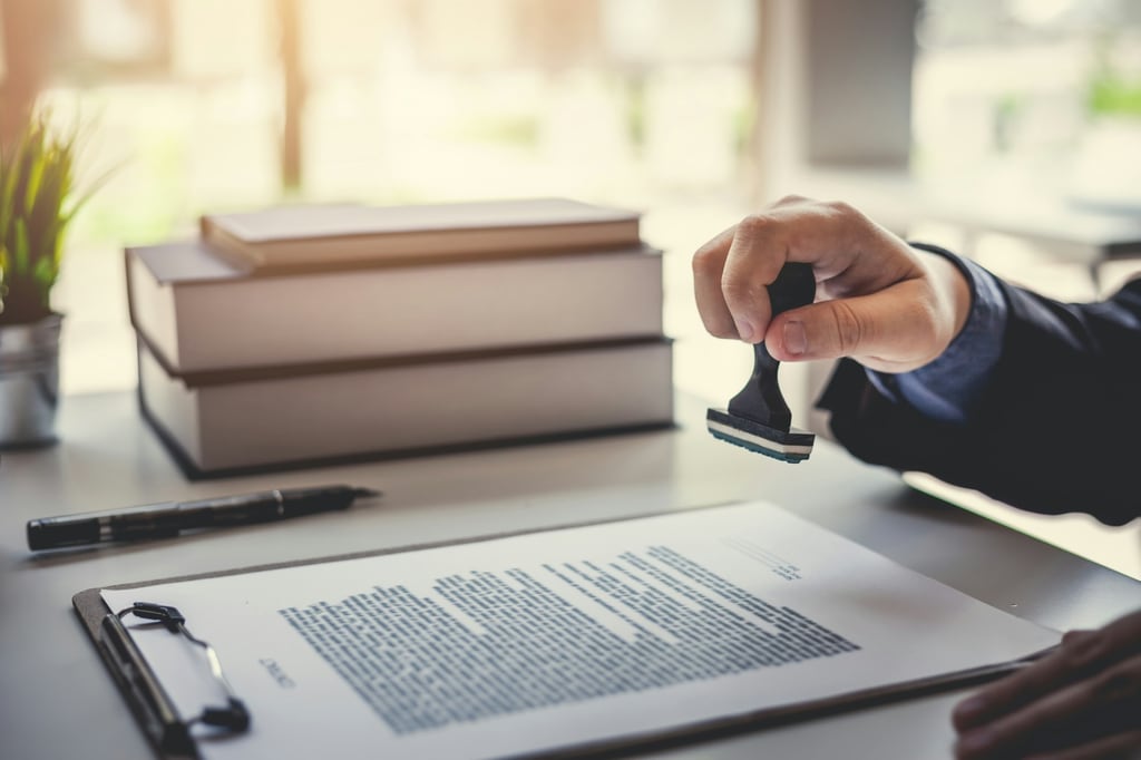 Main tenant un tampon encreur au-dessus d'un document juridique fixé sur une planchette à pince, avec une pile de livres et un stylo posés sur un bureau en arrière-plan