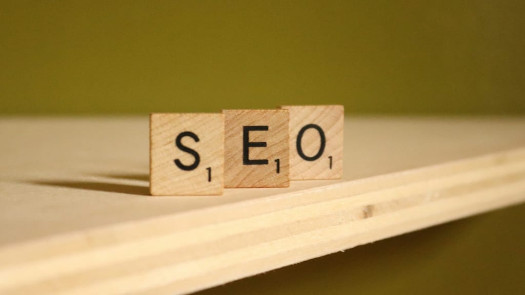 Three wooden Scrabble tiles spelling "SEO" resting on a light wooden surface against an olive green background.